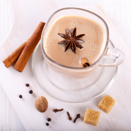 image shows a glass cup and saucer containing a chai drink with variety of spices surrounding and a star anise floating on the chai